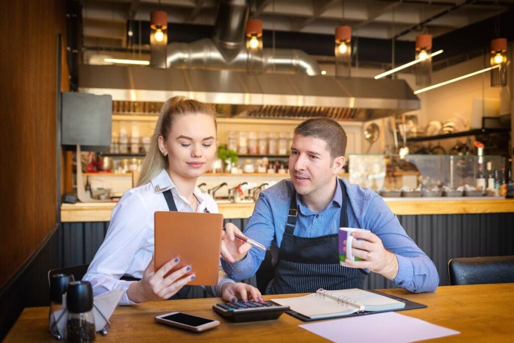 Dono e funcionária de restaurante conferindo números em um tablet, com calculadora e agenda sobre a mesa